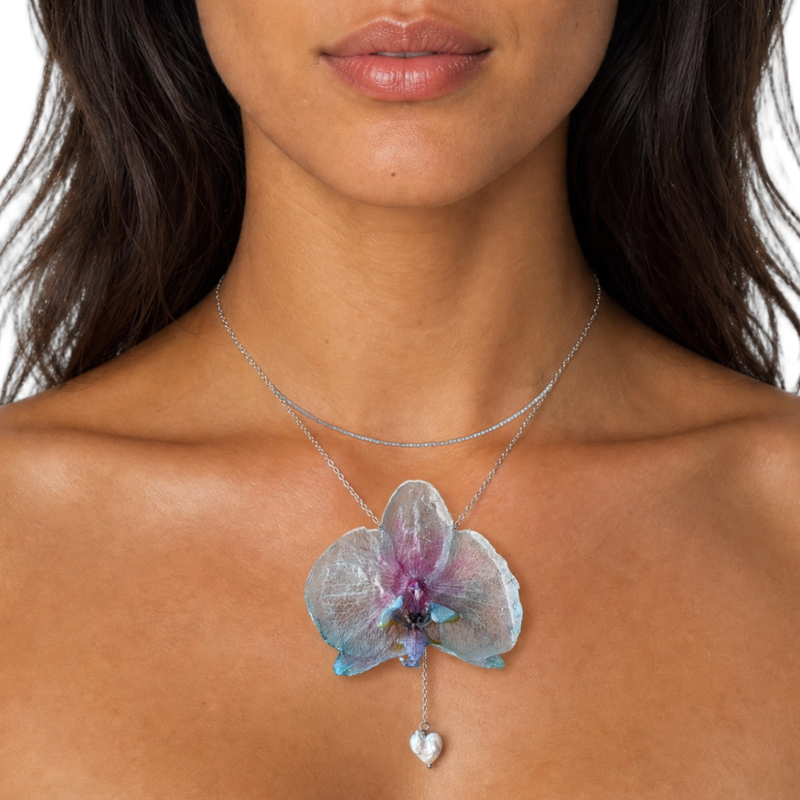 A woman modeling a delicate layered necklace featuring a translucent purple and blue orchid flower pendant with a small silver heart dangle, set against a solid white background.